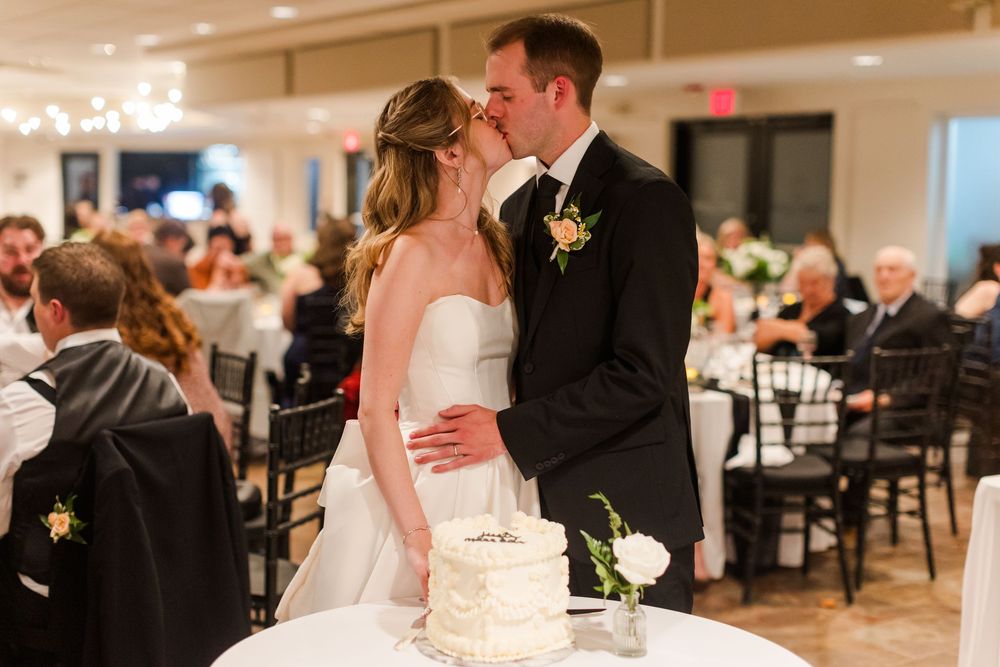 Bride and groom share a kiss at their wedding reception beside a white tiered cake in an elegant banquet hall.