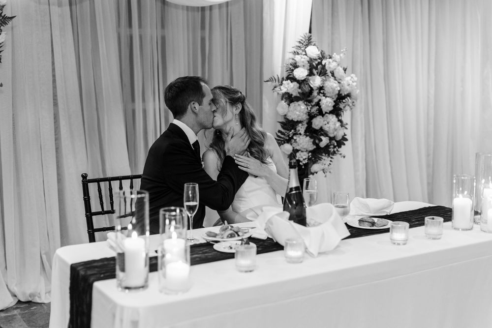 Bride and groom share a kiss at sweetheart table with candles and floral arrangement in black and white.