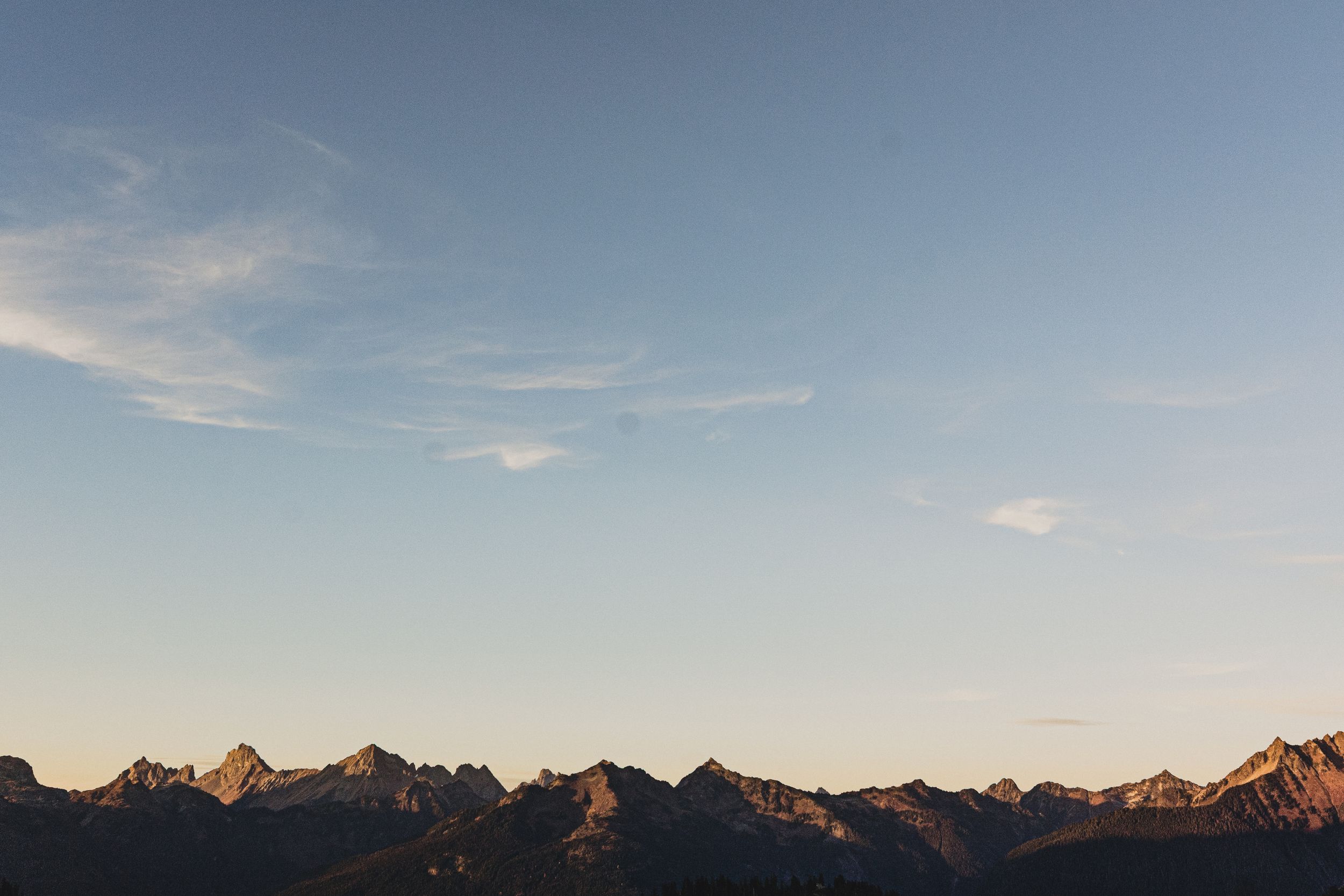 Jagged mountain peaks silhouetted at sunset beneath a soft blue sky with thin wispy clouds and a warm golden glow along the horizon.