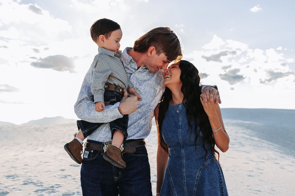 Family of three sharing a joyful moment on sandy dunes, father holding toddler while couple laughs together outdoors.