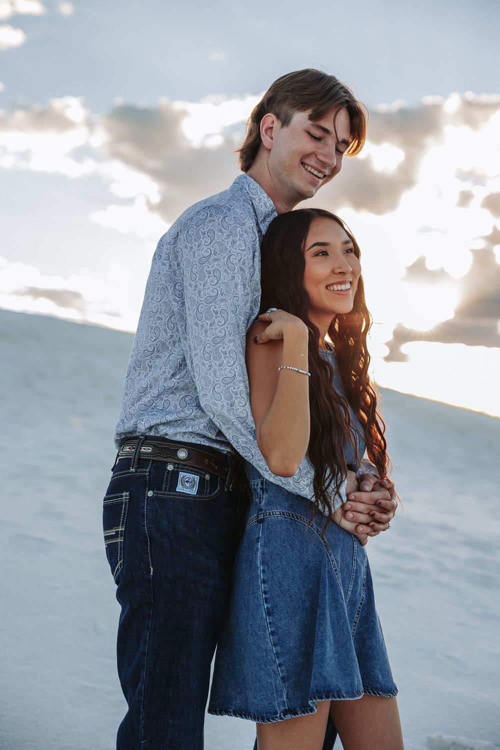 A smiling couple embraces on a sandy beach at sunset, woman in denim dress, man in gray shirt and jeans.