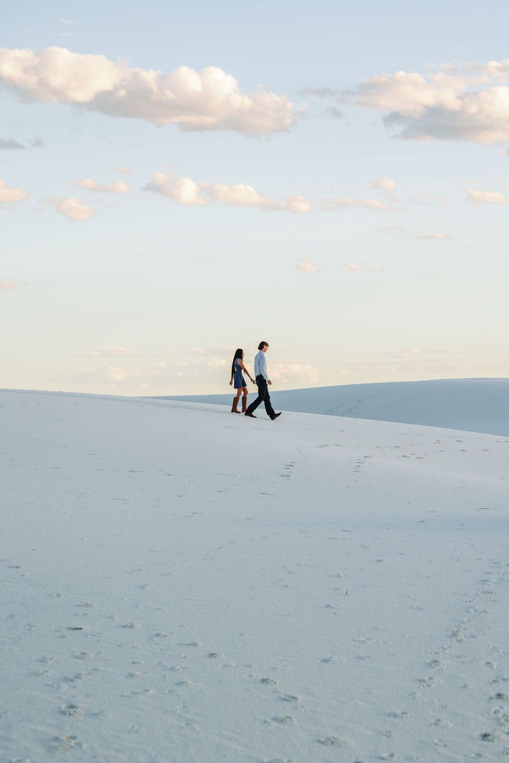 Couple walking hand in hand across white sand dunes under a partly cloudy sky at White Sands National Park.