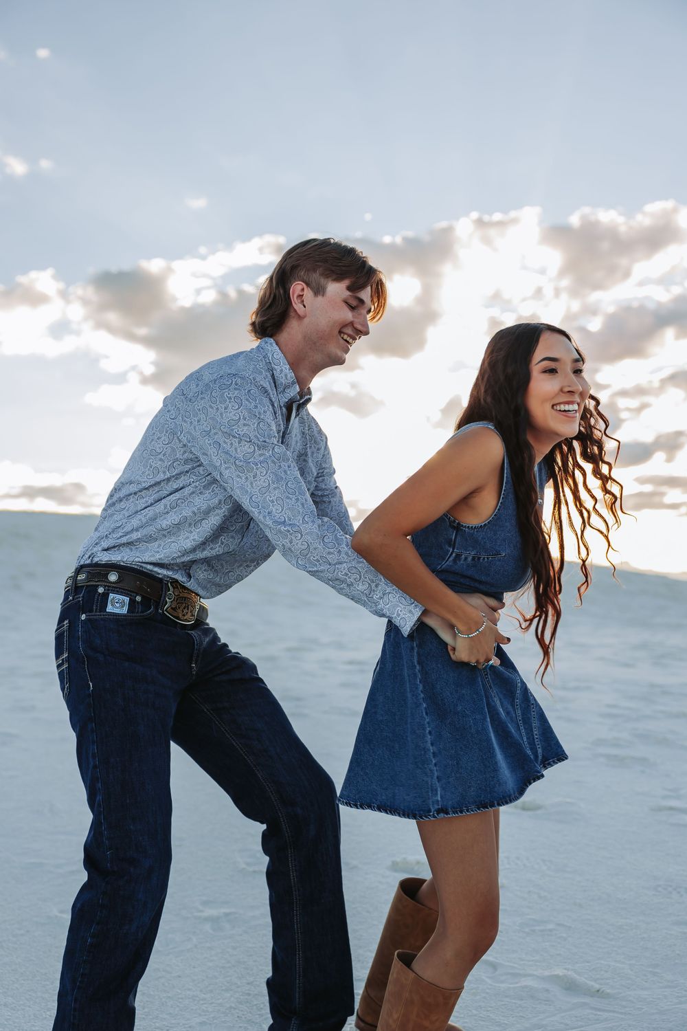 A laughing couple holds hands on white sand dunes, woman in denim dress and boots, man in floral shirt and jeans.