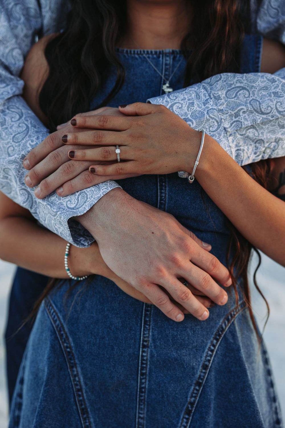 Close-up of couple embracing, showing hands with engagement ring, bracelets, and dark nail polish on denim outfit.