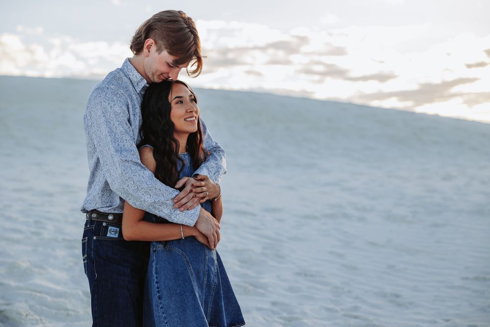 A couple embraces romantically on white sand dunes, the woman smiling in a blue dress while the man holds her close.
