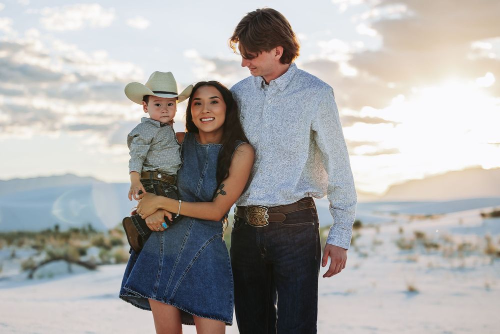 A young family poses outdoors at White Sands, with a toddler in a cowboy hat, mom in denim dress, and dad in a button-up shirt.