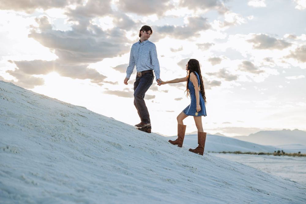 Couple holding hands walking up white sand dunes at White Sands National Park during golden hour.