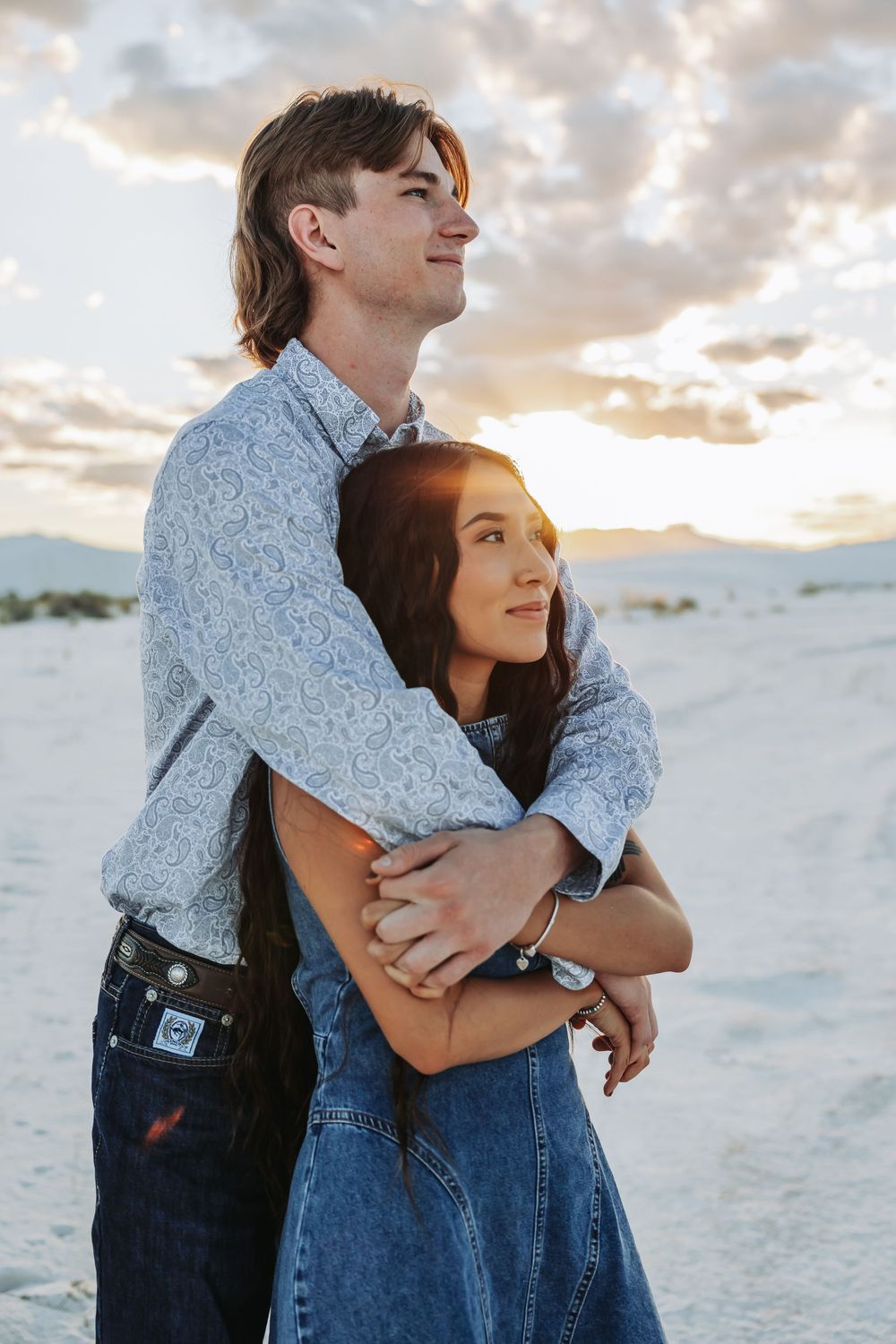 Couple embracing at sunset in white sand dunes, man wrapping arms around woman in denim dress.