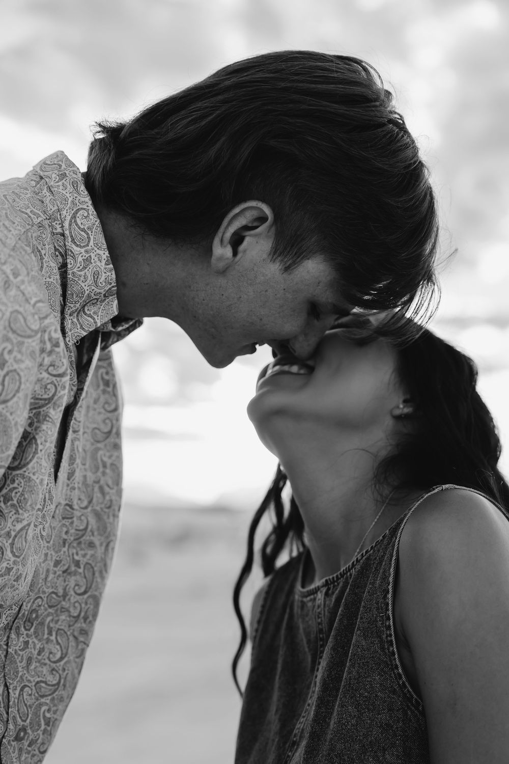 Black and white photo of a romantic couple sharing a tender moment, nearly kissing outdoors with cloudy sky background.