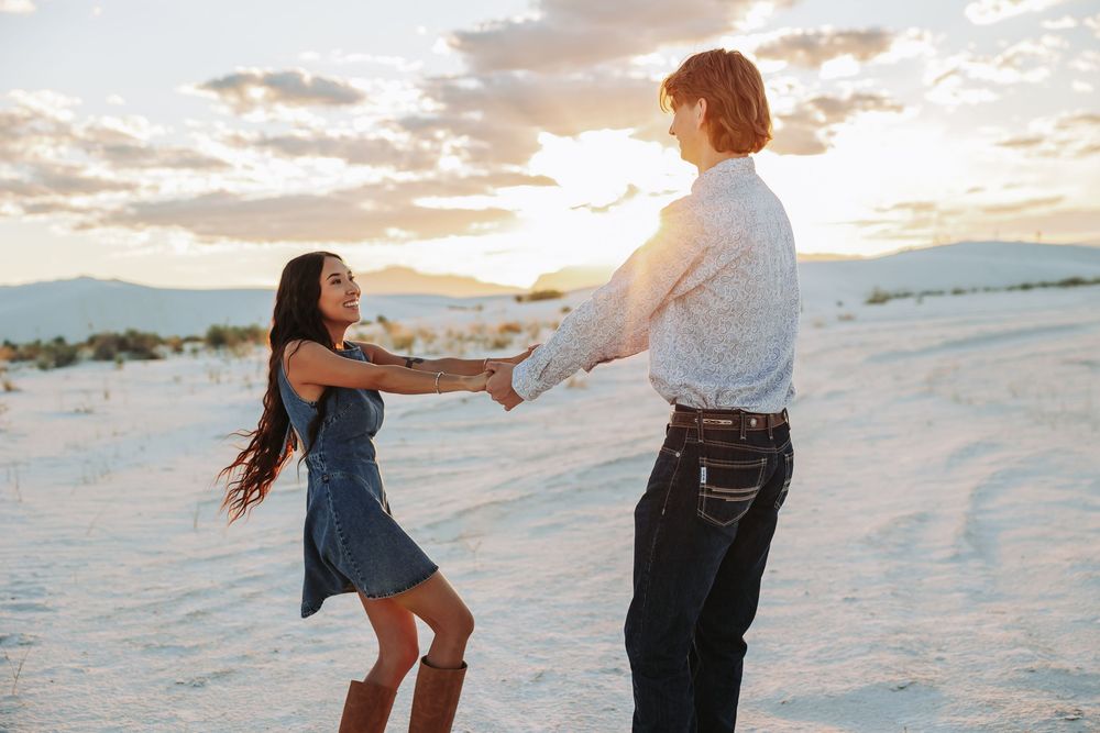 Couple dancing and holding hands on white sand dunes at sunset in a romantic outdoor setting.