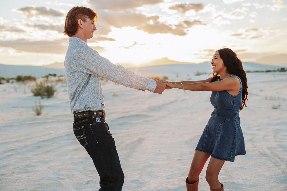 A couple holds hands and dances joyfully in a white sand desert at sunset.