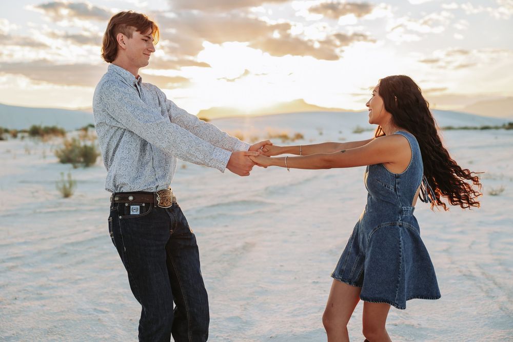 Couple dancing and holding hands on white sand dunes at sunset in a romantic outdoor setting.