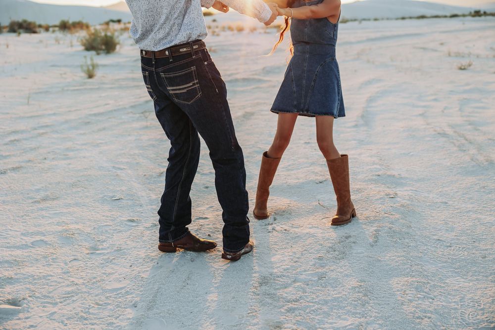 Couple dancing in cowboy boots on white sand desert, man in dark jeans and woman in denim skirt.