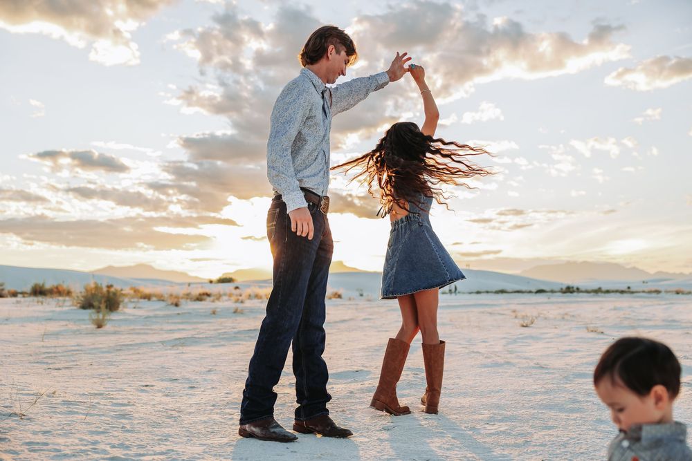 Couple dancing together in white sand desert at sunset, man spinning woman in blue dress with child nearby.