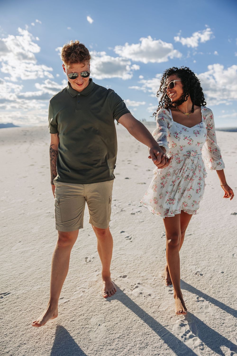 A happy interracial couple holds hands while walking barefoot on white sand dunes under a partly cloudy blue sky.