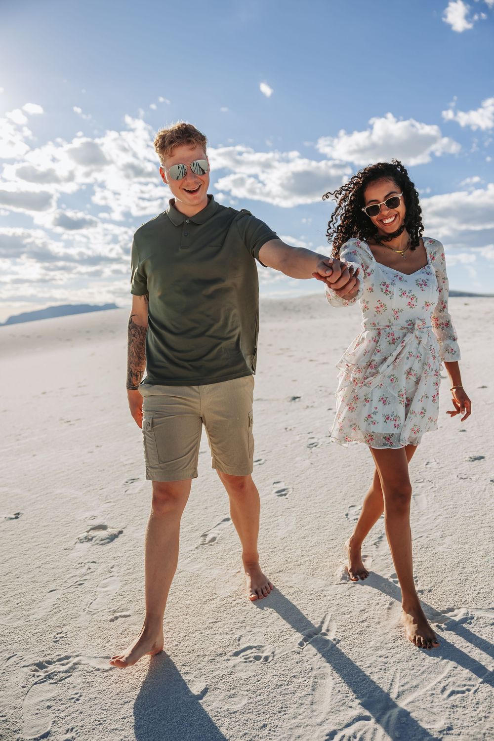 Happy interracial couple holding hands walking barefoot on white sand beach on a sunny day.
