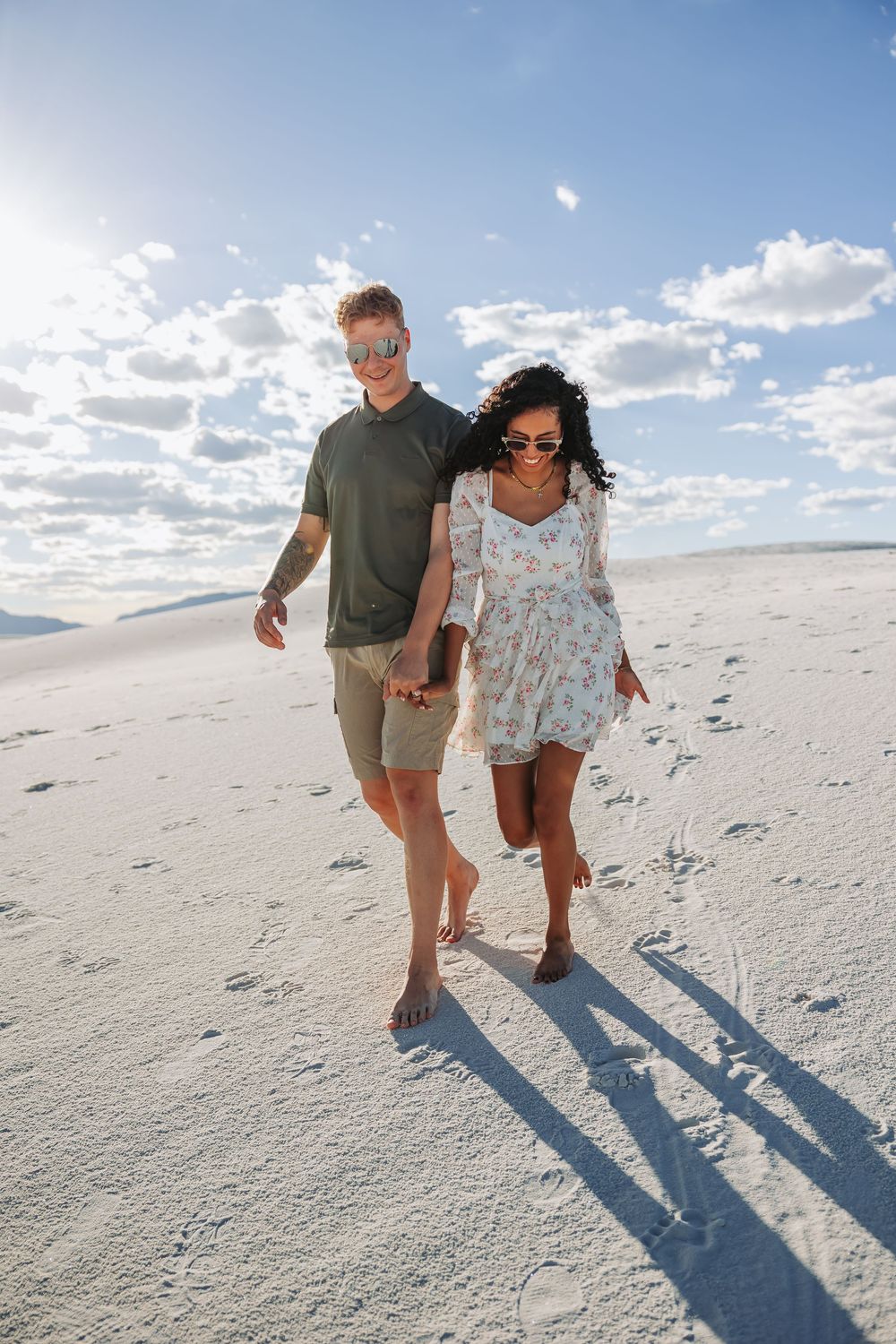 A couple walks barefoot hand-in-hand across white sand dunes under a bright blue sky with scattered clouds.