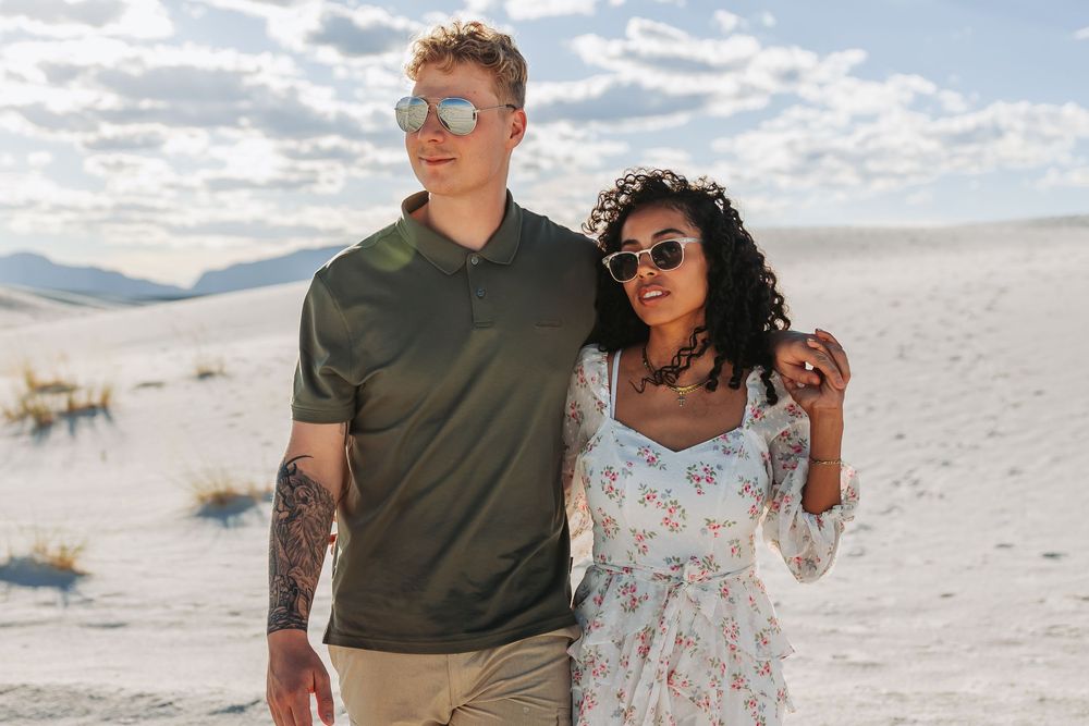 A couple walks together on white sand dunes, man with tattoos in green polo, woman in floral dress, both wearing sunglasses.