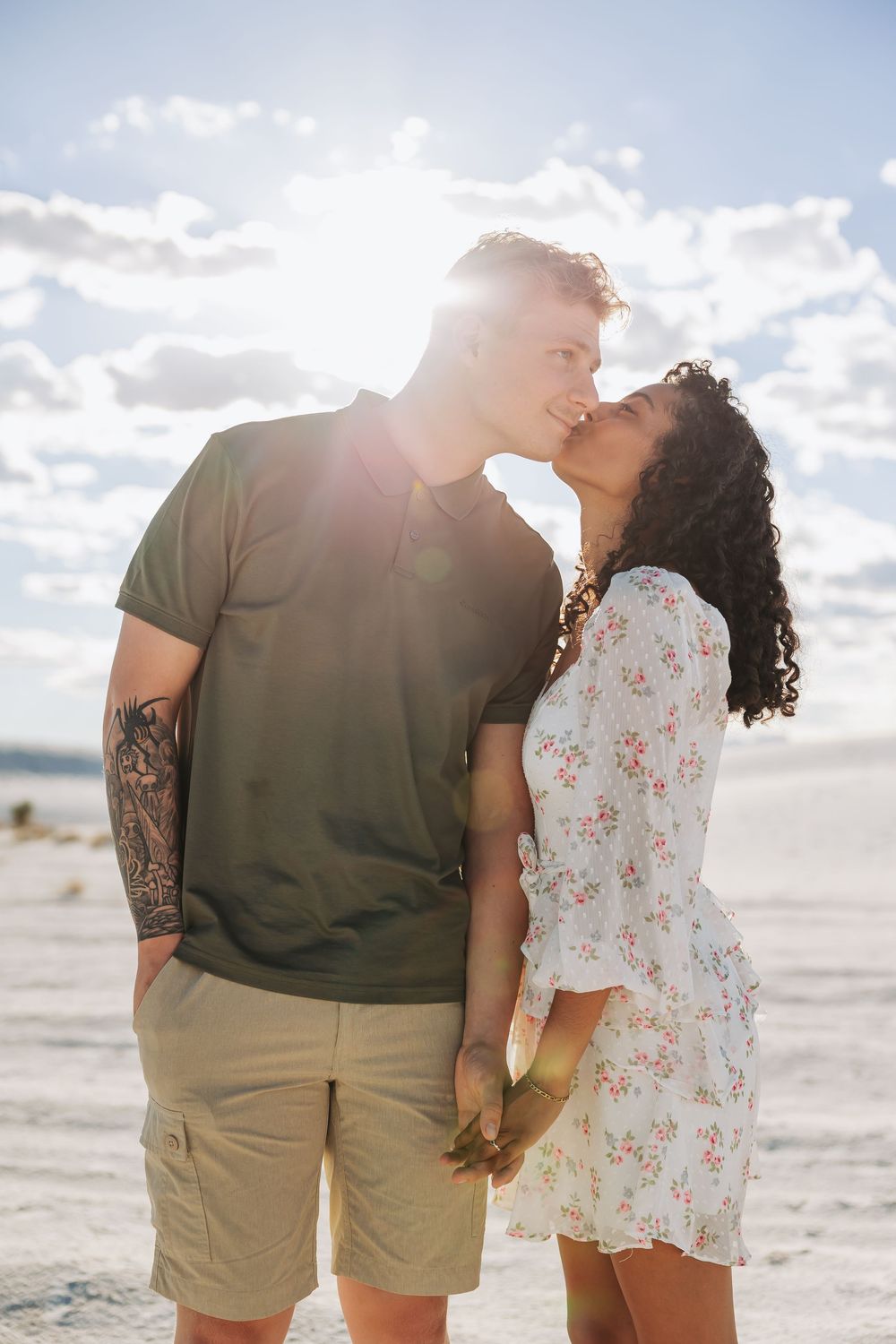 A couple sharing a romantic kiss while holding hands on a sunny beach, man with tattoos in green shirt and woman in floral dress.