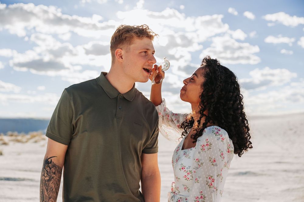 A young interracial couple shares a playful moment outdoors on a sandy beach under a cloudy sky.