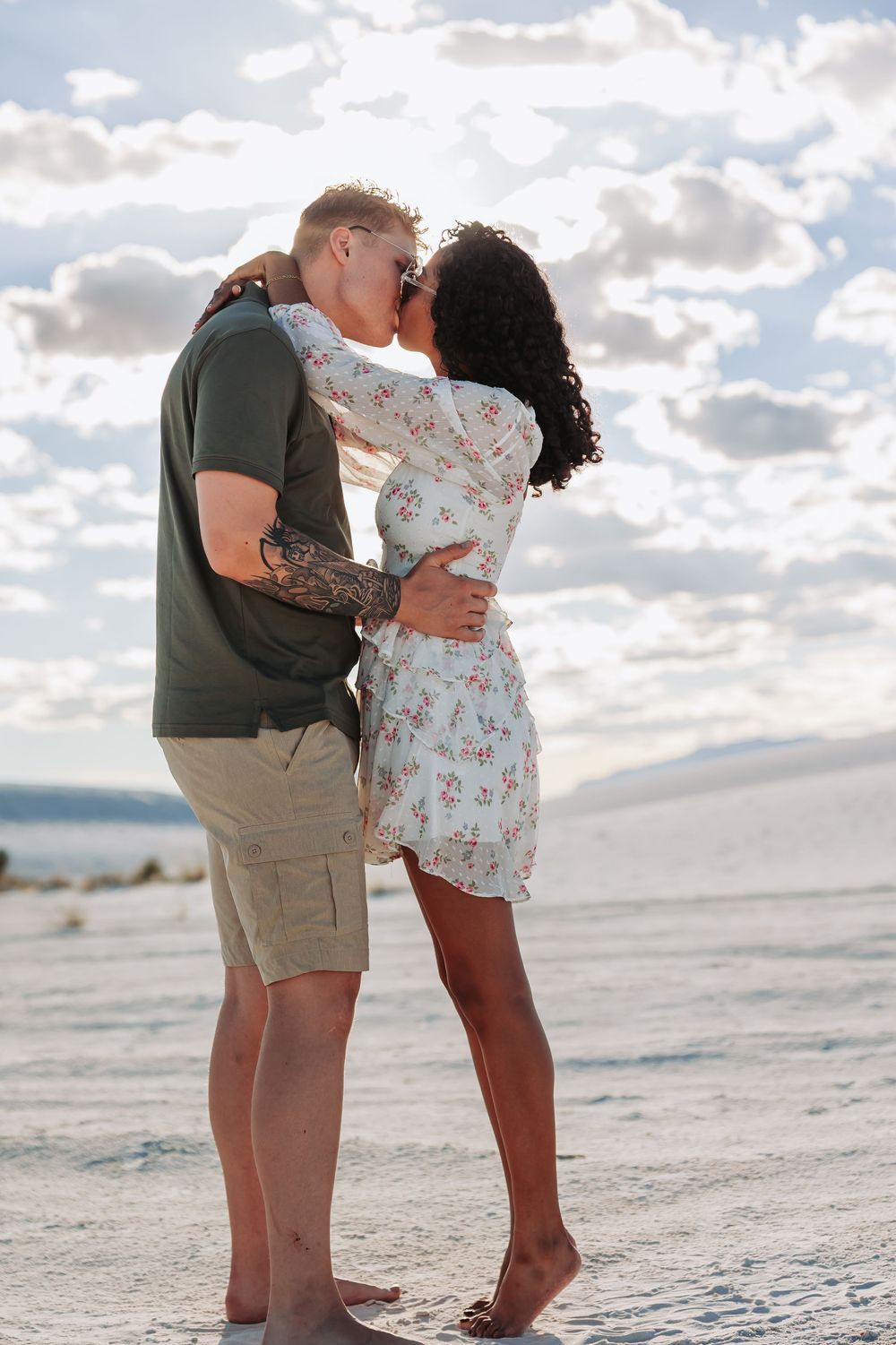 Interracial couple sharing a romantic kiss on white sand dunes under a cloudy sky, woman in floral dress.