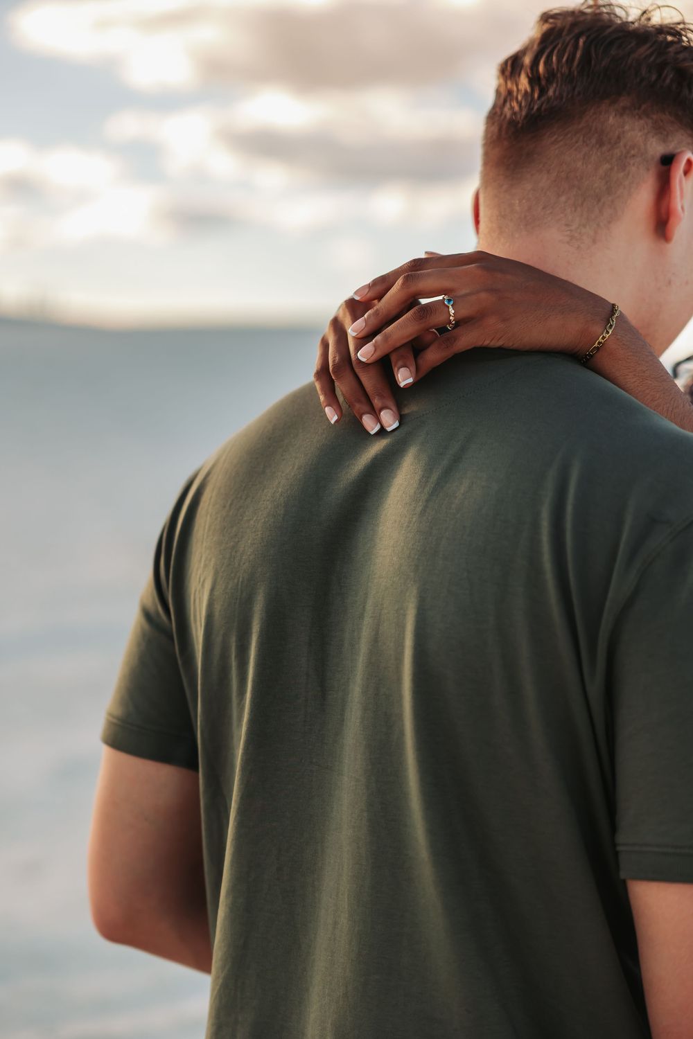 A woman's hand with rings rests on a man's shoulder in a green t-shirt, overlooking a serene ocean view.