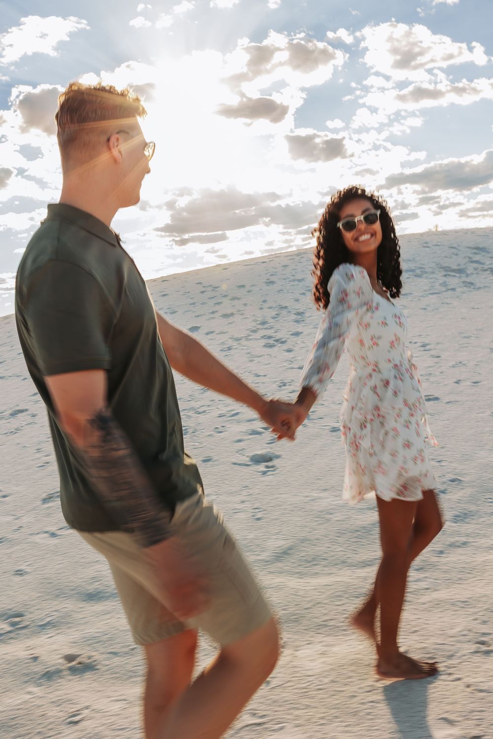 Couple holding hands walking on sandy beach, woman in floral dress smiling, man in dark shirt, sunny sky backdrop.
