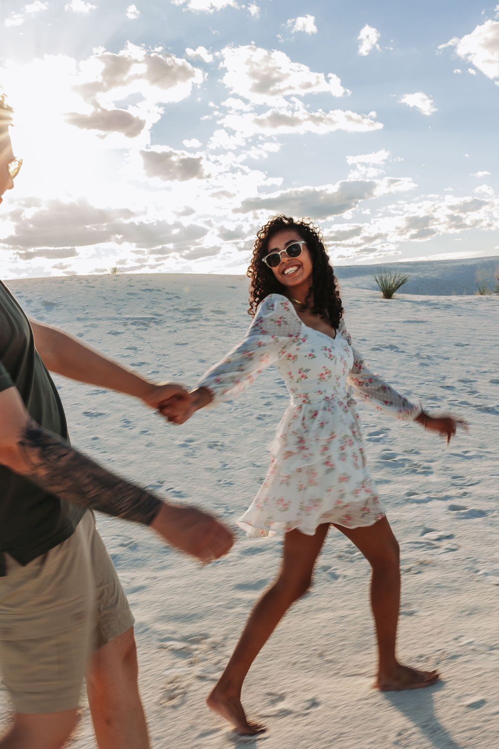 A joyful woman in a floral dress holds hands with a partner while walking barefoot on white sand dunes.