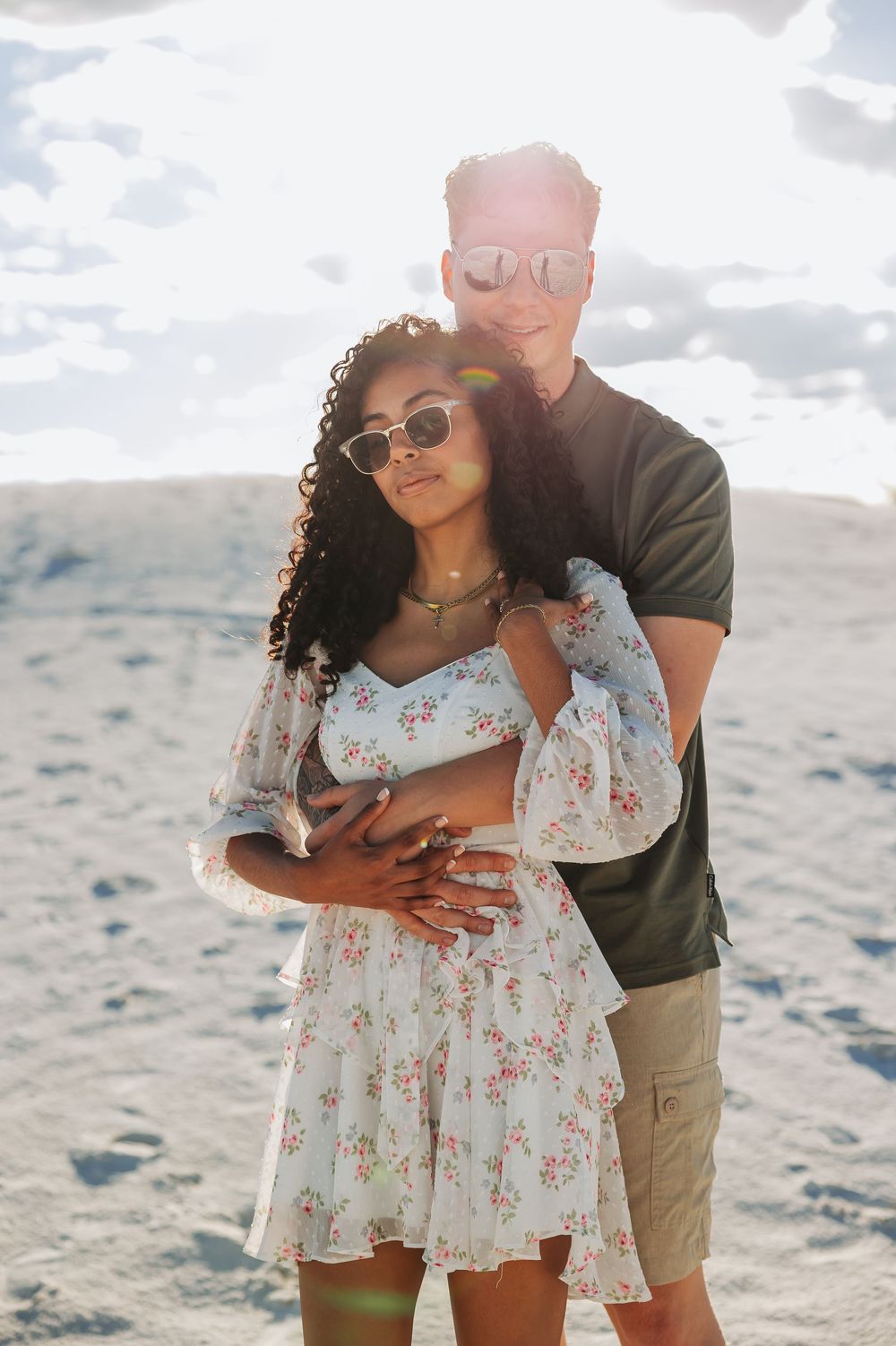 A couple embraces on sandy dunes, woman in floral dress and sunglasses, man in olive shirt, backlit by bright sun.