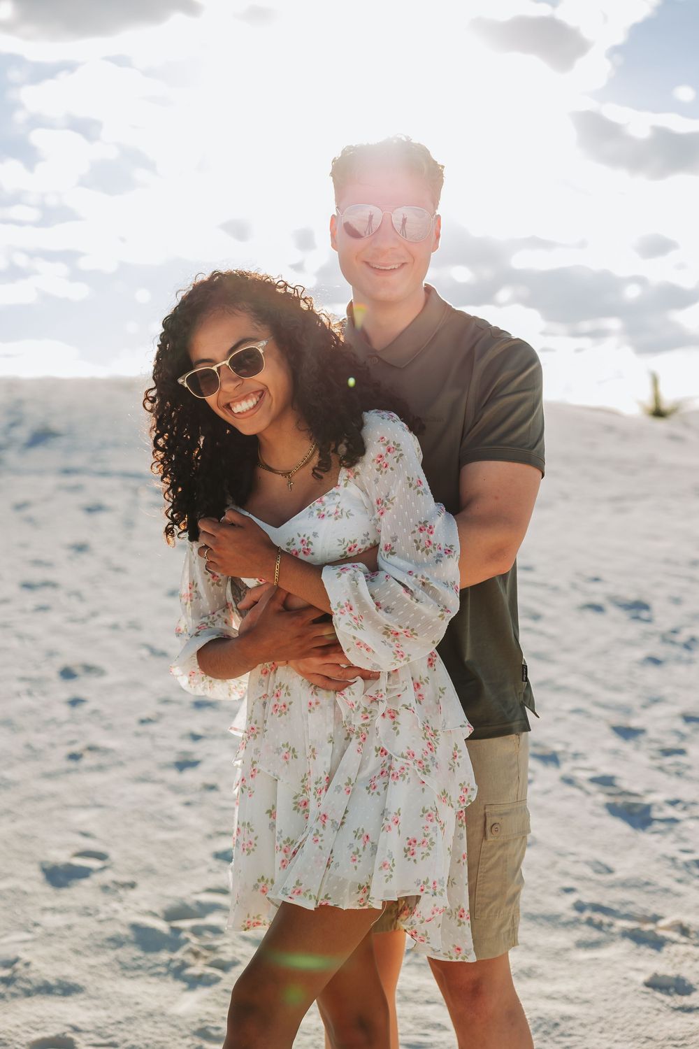 A happy interracial couple embraces on a sandy beach, woman in floral dress and sunglasses, man in dark polo shirt.