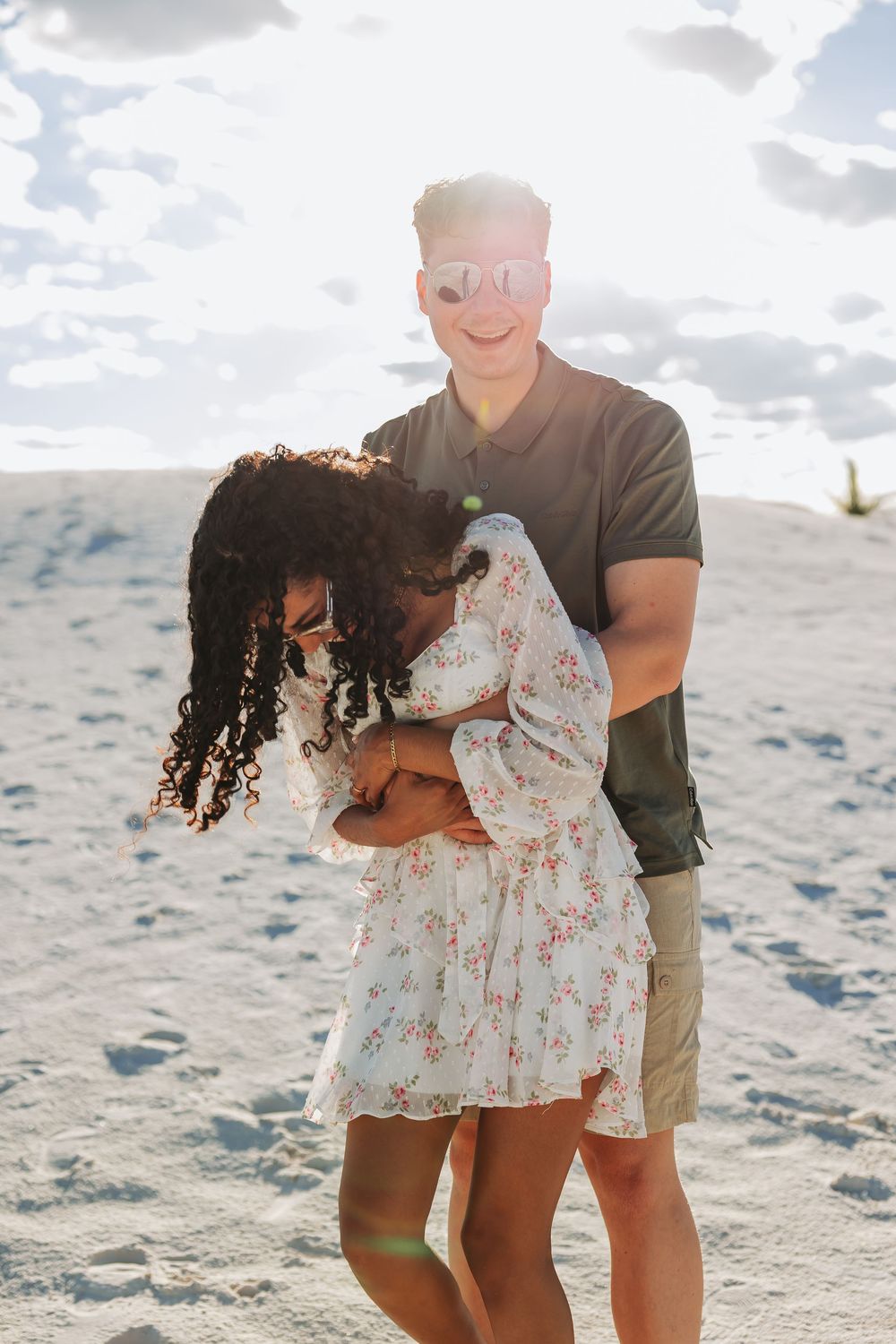 A happy interracial couple embracing on a sandy beach, backlit by bright sunlight, woman in floral dress, man in dark polo.