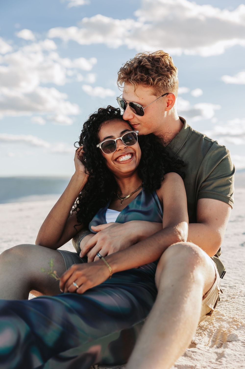 Happy interracial couple sitting on sandy beach, woman in sunglasses smiling as man embraces her under cloudy sky.