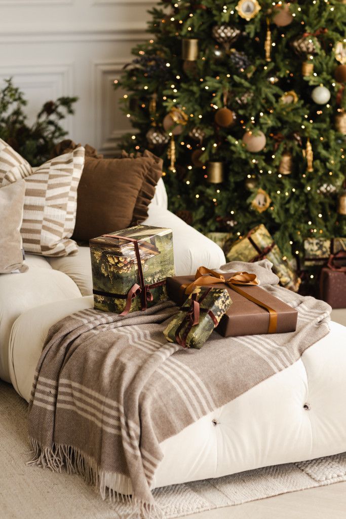 Cozy Christmas living room with a cream sofa, velvet pillows, wooden nesting tables, gift wrap, and a garland-draped fireplace.