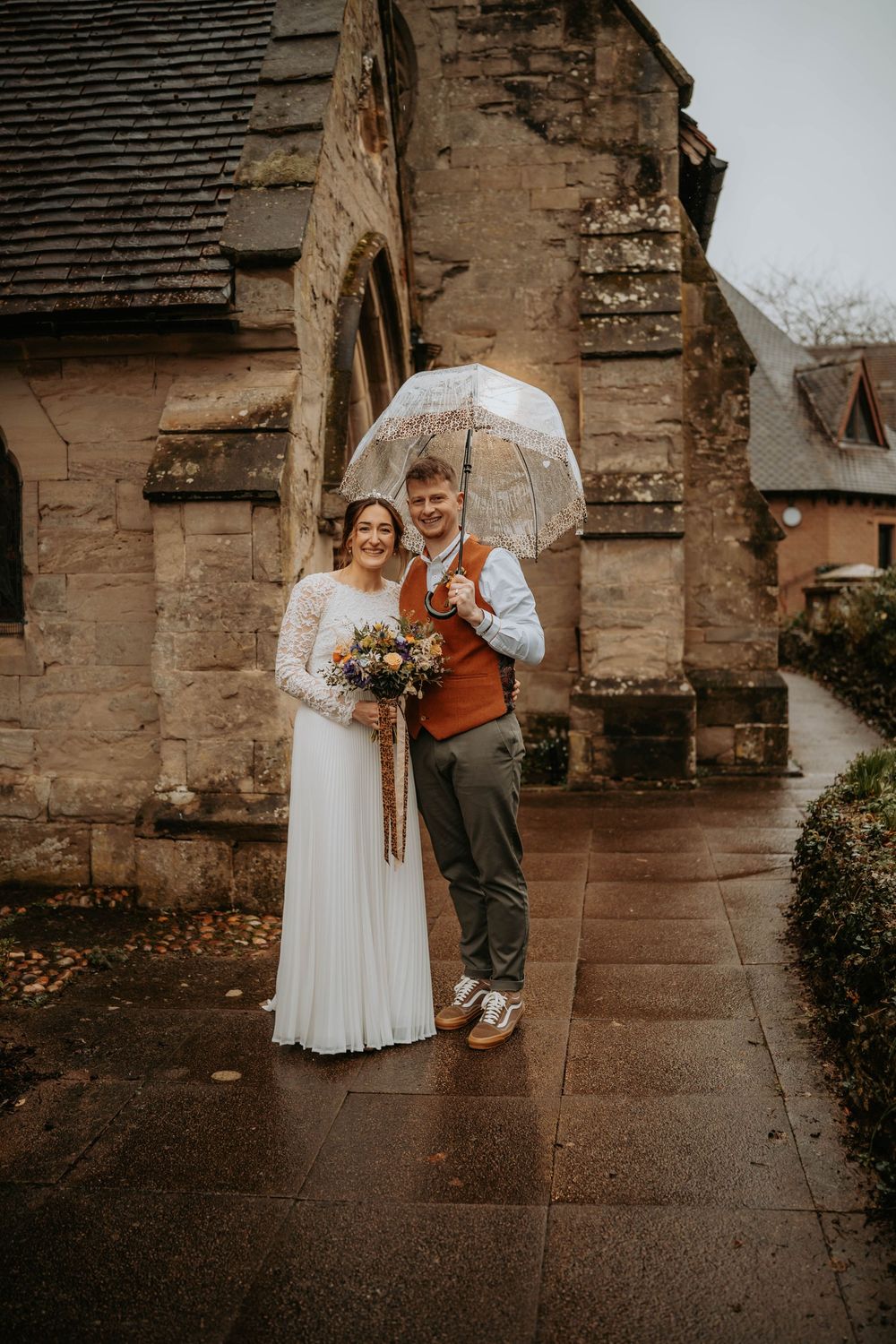 Bride and groom share umbrella outside stone church on rainy wedding day.