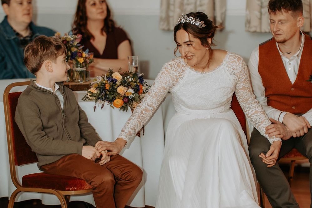 Bride in white lace gown holds hands with young boy in brown suit at wedding ceremony.