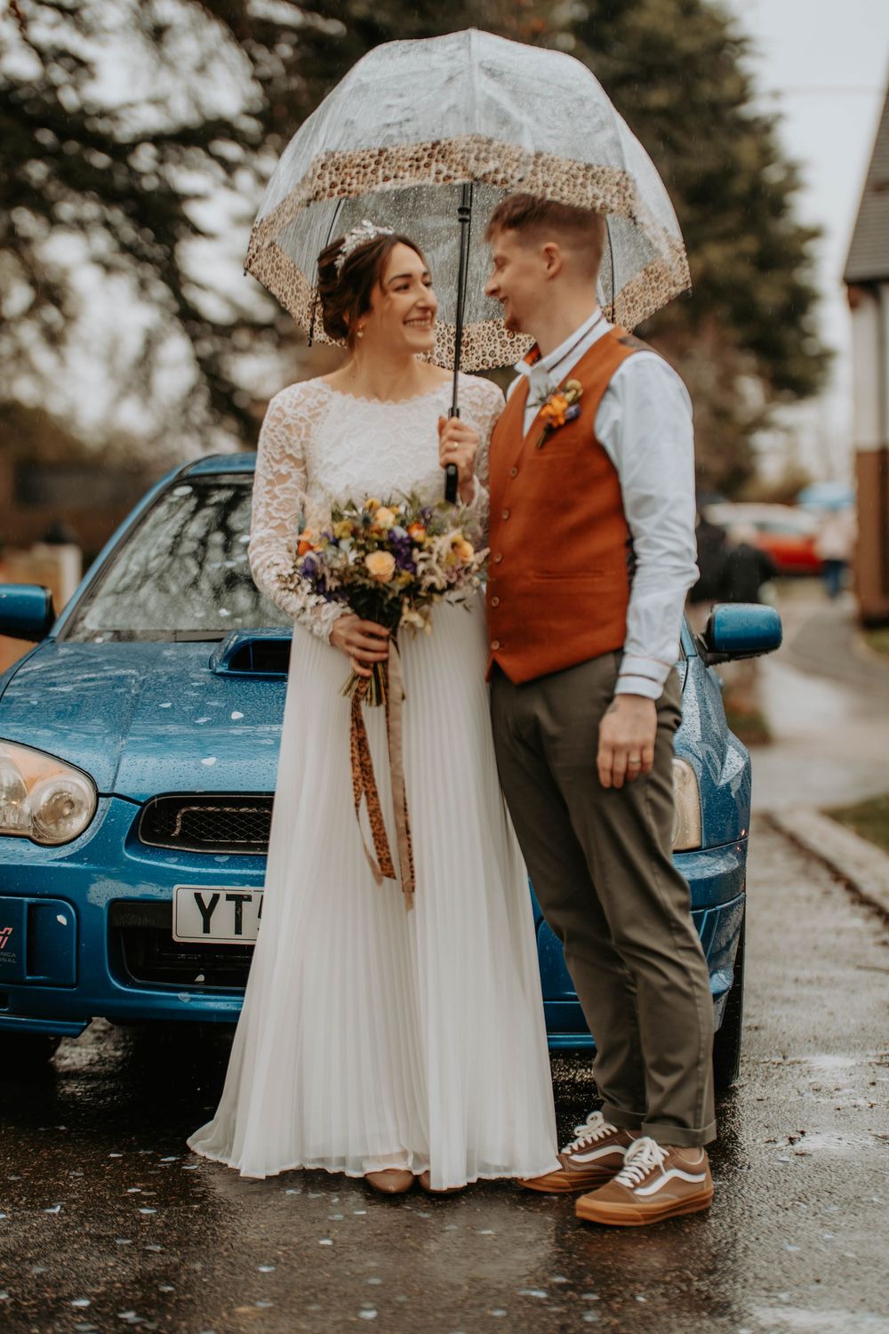 Bride and groom sharing umbrella in rain beside blue car, bride holding wildflower bouquet in white dress.