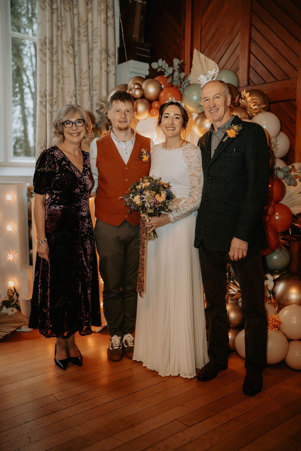 Wedding party of four posing before balloon arch decor, bride in white gown holding bouquet, groom in rust vest.