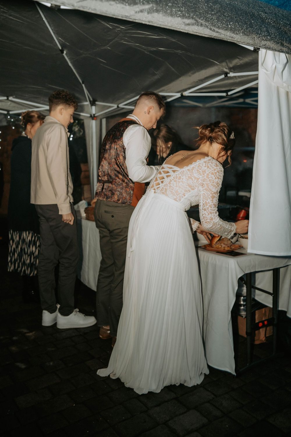 Bride in lace wedding dress at outdoor food station with guests during evening wedding reception.