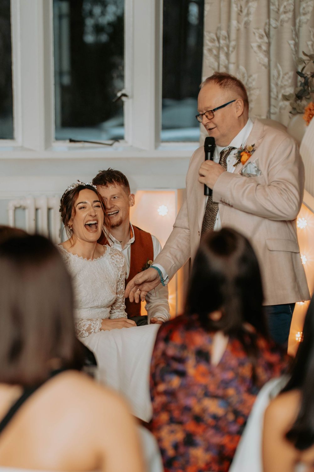 Bride and groom laugh joyfully as a man in a beige blazer gives a wedding speech with a microphone.