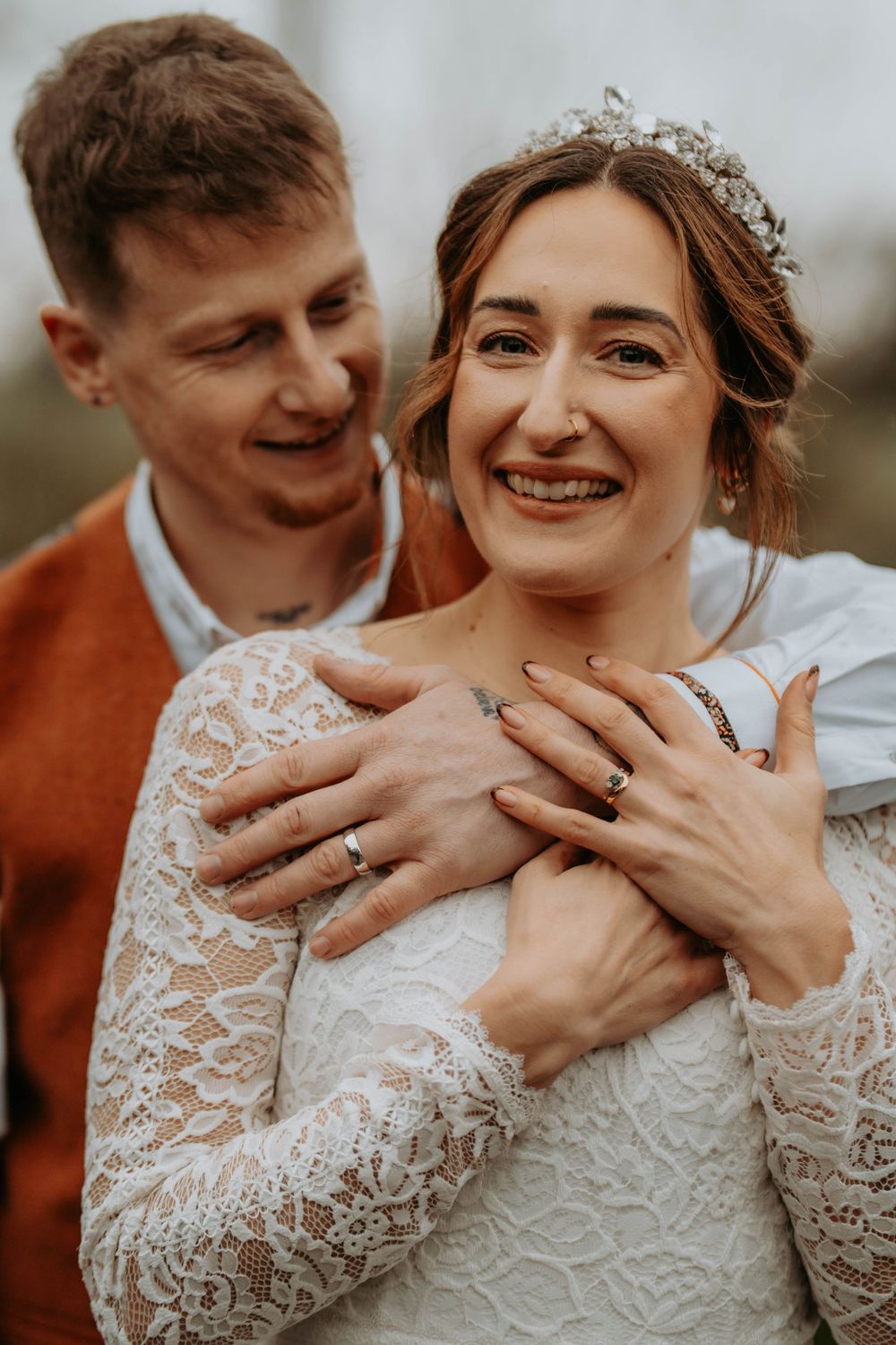 Groom embraces smiling bride wearing lace dress and tiara at wedding.