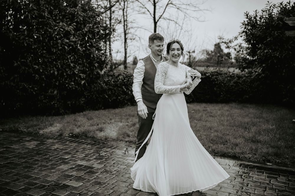 Black and white photo of a bride and groom posing outdoors on a brick path surrounded by trees.