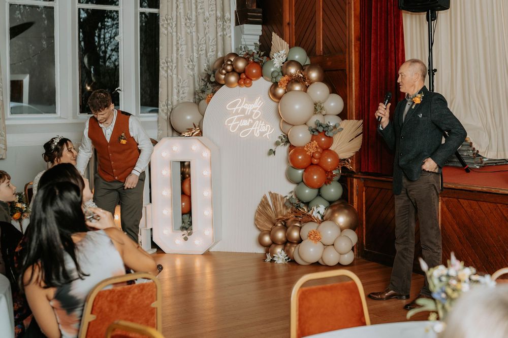 Elegant party setup with balloon arch in terracotta and sage, marquee number 0, and guests gathered in decorated hall.