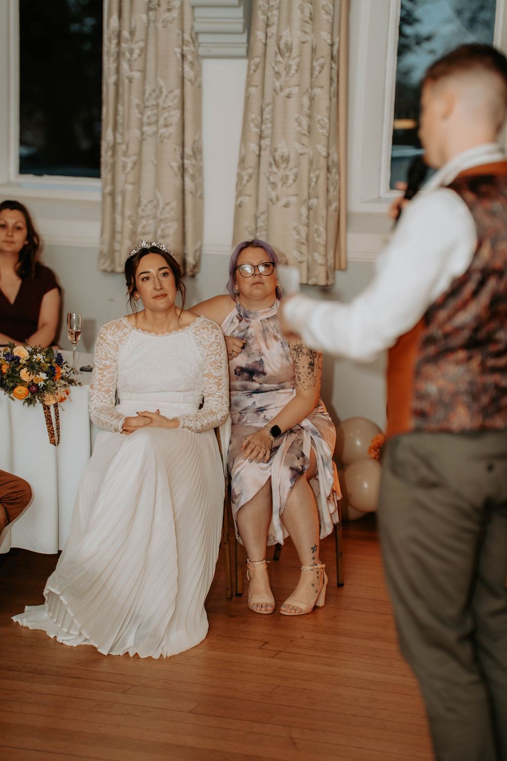 Bride in lace wedding dress seated with guest during indoor wedding reception speech.