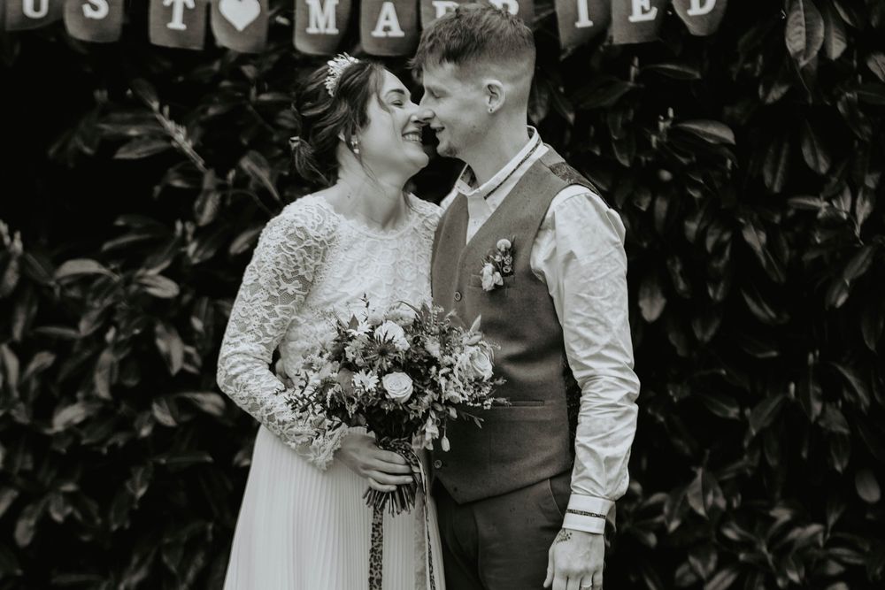 Black and white photo of newlyweds sharing a romantic moment under a Just Married banner holding a bridal bouquet.
