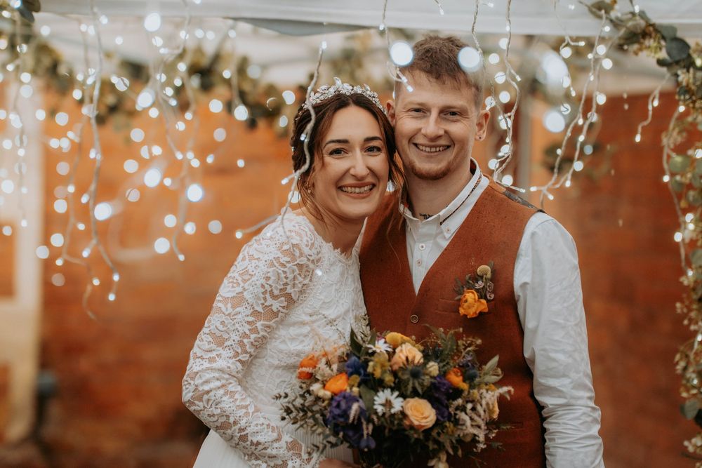 Happy bride and groom smiling together under fairy lights, bride holding colorful bouquet at rustic wedding venue.