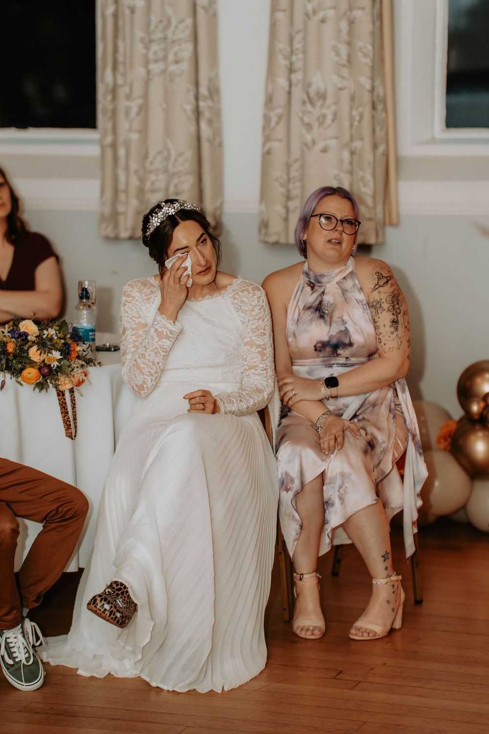 Emotional bride wiping tears while seated next to guest during wedding reception speech.