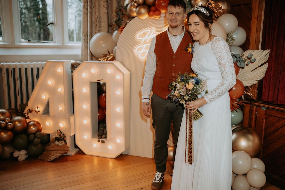Couple in wedding attire posing beside illuminated 40 marquee letters and balloon arch decorations.