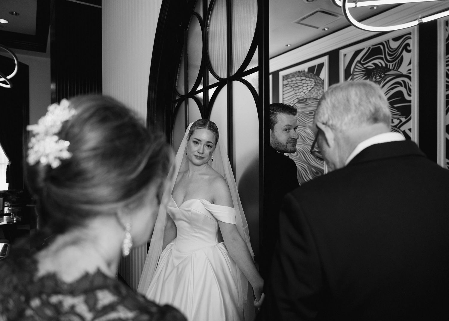 Black and white photo of a bride in a white gown and veil speaking with guests at an elegant wedding venue.