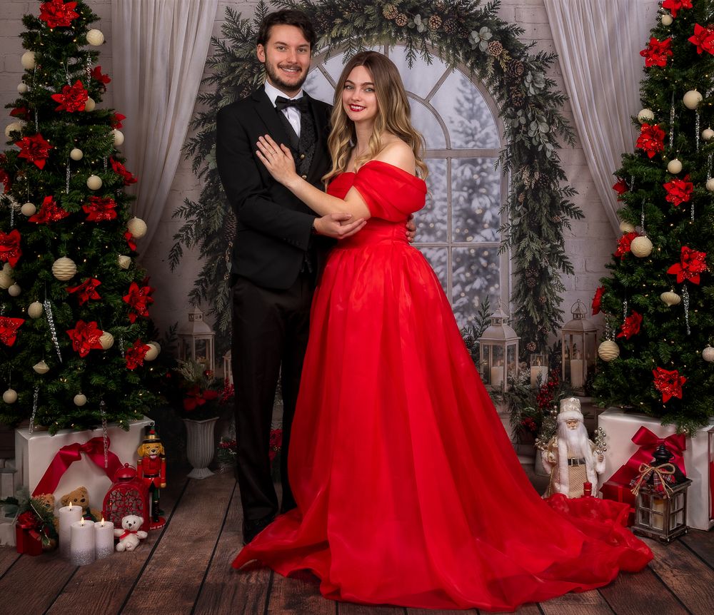Couple in a black tuxedo and flowing red off-the-shoulder gown pose in a festive Christmas studio with decorated trees and gifts.