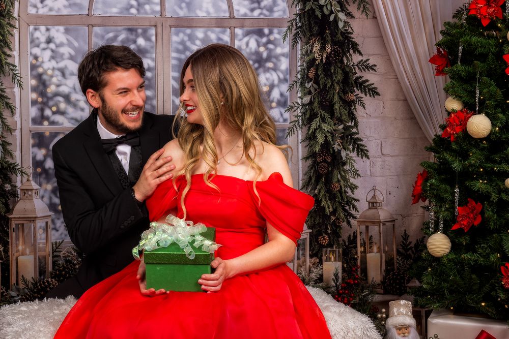 Smiling man in a black tuxedo embraces a woman in a red off-shoulder dress holding a green Christmas gift beside a decorated tree.